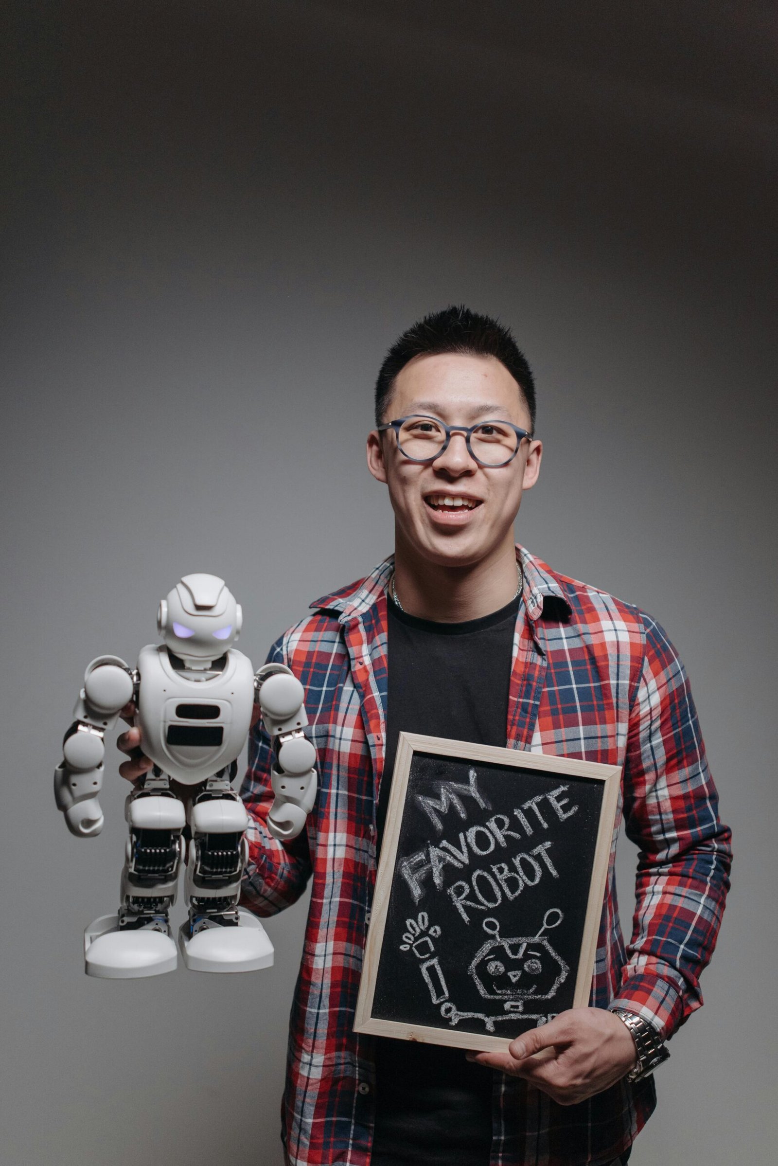 Asian man in glasses holding a toy robot and chalkboard in a studio setting.